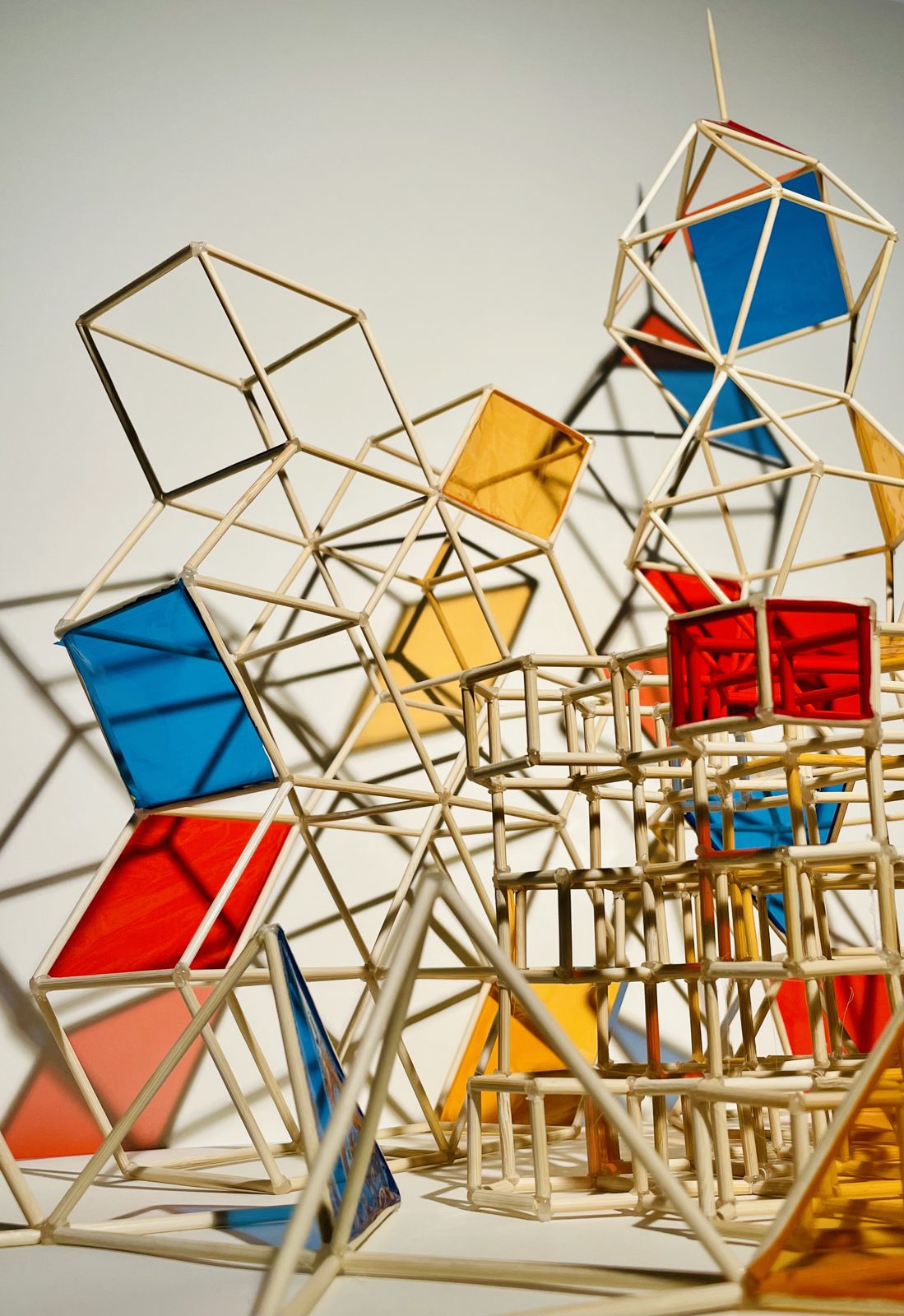 A sculptural model of a city made from a lattice of thin wooden sticks, with translucent blue, red and yellow panels casting coloured shadows